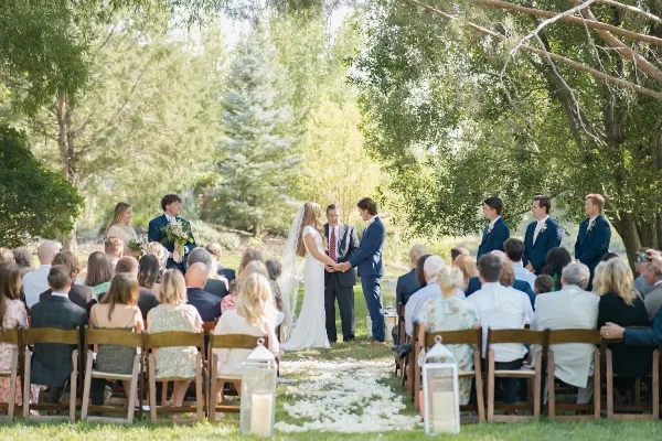 Outdoor wedding ceremony with bride and groom exchanging vows under trees, surrounded by seated guests.