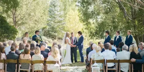 Outdoor wedding ceremony with bride and groom exchanging vows under trees, surrounded by seated guests.