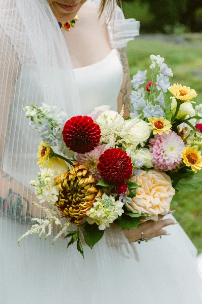 A bride in a white dress holding a large, colorful bouquet featuring red dahlias, yellow zinnias, and white flowers.