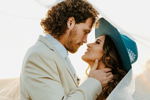 A close-up, warm-toned photograph of a groom in a light suit and a bride in a teal hat and veil, gazing at each other affectionately.