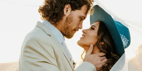 A close-up, warm-toned photograph of a groom in a light suit and a bride in a teal hat and veil, gazing at each other affectionately.