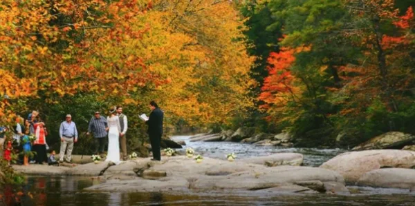 A wedding ceremony taking place outdoors on rocks beside a stream, with an officiant, bride, groom, and guests present.