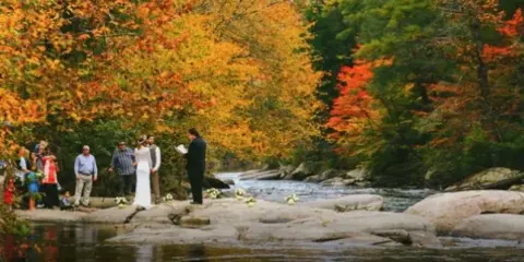 A wedding ceremony taking place outdoors on rocks beside a stream, with an officiant, bride, groom, and guests present.
