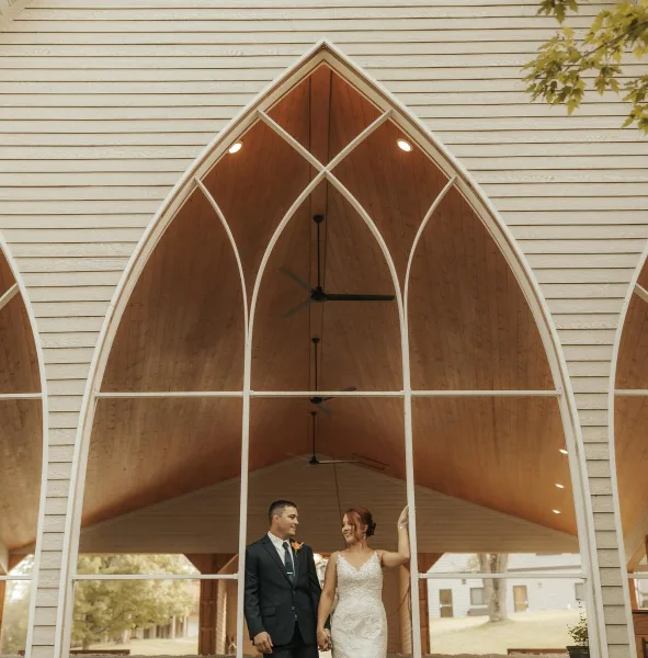 A bride and groom standing inside a large, open-air chapel structure with tall arched windows and a wooden ceiling.