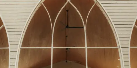 A bride and groom standing inside a large, open-air chapel structure with tall arched windows and a wooden ceiling.