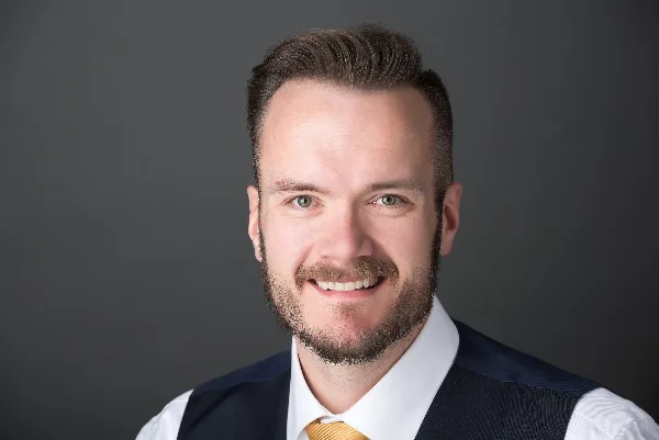 Professional headshot of a smiling man with a beard, wearing a white shirt, dark vest, and gold tie.