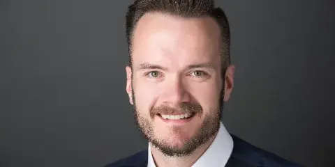 Professional headshot of a smiling man with a beard, wearing a white shirt, dark vest, and gold tie.