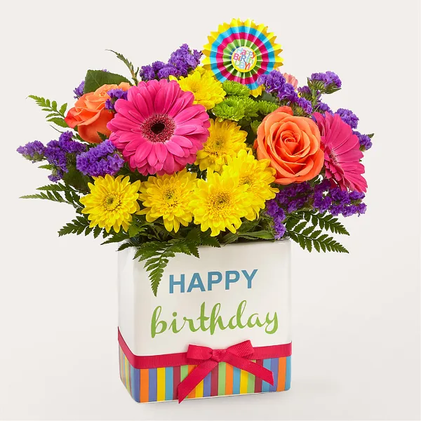 A colorful floral arrangement featuring pink gerbera daisies, yellow chrysanthemums, and orange roses in a white container with a 'Happy Birthday' message.