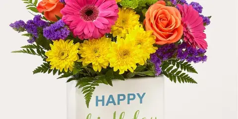 A colorful floral arrangement featuring pink gerbera daisies, yellow chrysanthemums, and orange roses in a white container with a 'Happy Birthday' message.