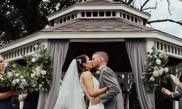 A bride and groom sharing a kiss at a white gazebo decorated with floral arrangements and grey drapes.