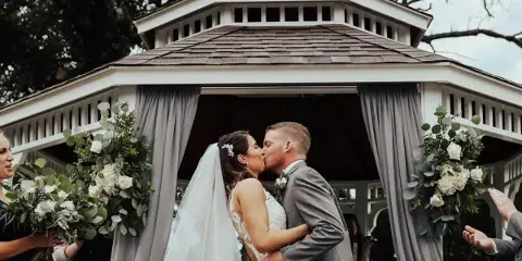 A bride and groom sharing a kiss at a white gazebo decorated with floral arrangements and grey drapes.