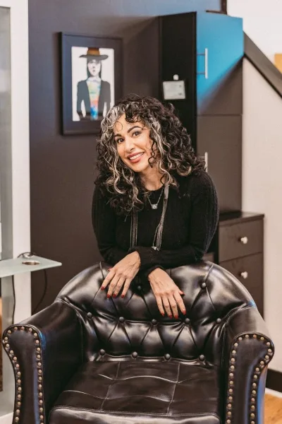 A smiling woman with curly highlighted hair seated in a black leather salon chair inside a hair studio.