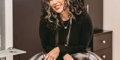 A smiling woman with curly highlighted hair seated in a black leather salon chair inside a hair studio.