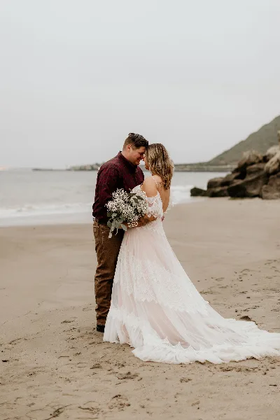 A bride and groom standing on a beach, the bride holding a bouquet of white flowers and wearing a long lace wedding dress.