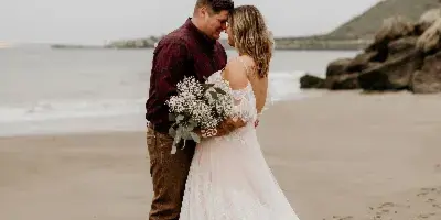 A bride and groom standing on a beach, the bride holding a bouquet of white flowers and wearing a long lace wedding dress.