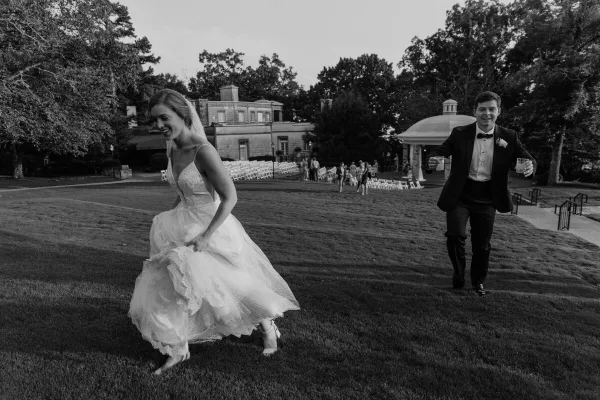 A black and white photo of a bride and groom running joyfully across a grassy lawn at a wedding venue.