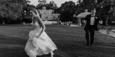 A black and white photo of a bride and groom running joyfully across a grassy lawn at a wedding venue.