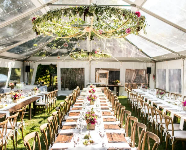Interior of a clear-frame wedding tent with long tables, wooden chairs, and floral centerpieces.