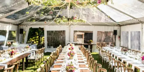 Interior of a clear-frame wedding tent with long tables, wooden chairs, and floral centerpieces.