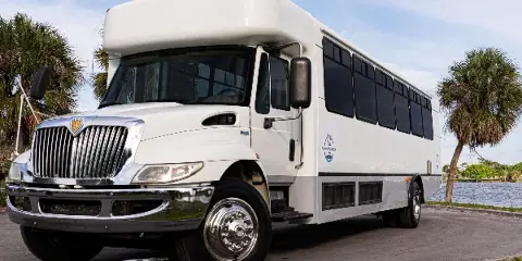 A white 32-passenger shuttle bus with the Performance logo parked near a waterfront with palm trees.