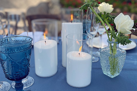 A wedding table setting featuring lit pillar candles, blue glassware, and white flowers in a glass vase.