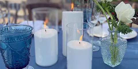 A wedding table setting featuring lit pillar candles, blue glassware, and white flowers in a glass vase.