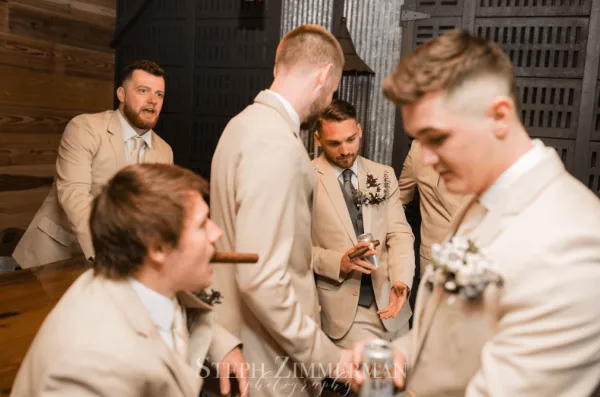 Groomsmen in beige suits socializing inside a rustic barn venue with wood and metal walls.