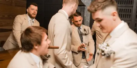 Groomsmen in beige suits socializing inside a rustic barn venue with wood and metal walls.
