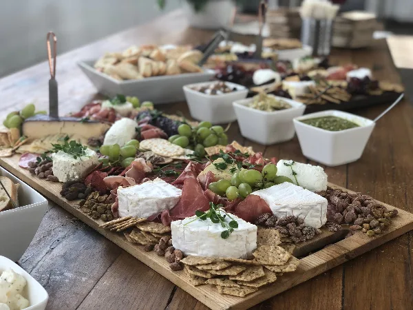 A wooden board filled with assorted cheeses, cured meats, grapes, crackers, and nuts, set on a table with additional catering bowls in the background.