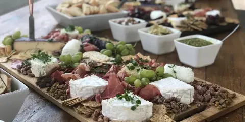 A wooden board filled with assorted cheeses, cured meats, grapes, crackers, and nuts, set on a table with additional catering bowls in the background.