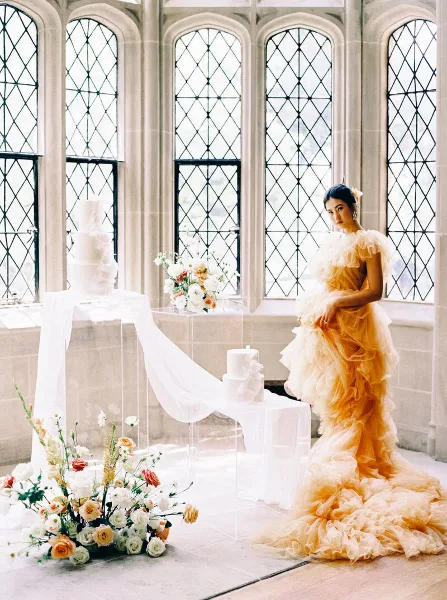 A bride in a ruffled peach gown stands next to a wedding cake and floral arrangements in a historic venue with large arched windows.