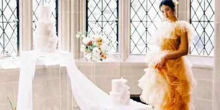 A bride in a ruffled peach gown stands next to a wedding cake and floral arrangements in a historic venue with large arched windows.