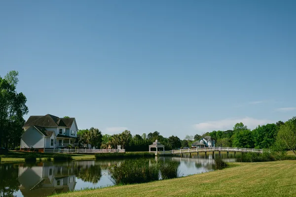 A scenic view of a lakefront wedding venue featuring a white bridge, a gazebo, and houses surrounded by green lawns and trees.