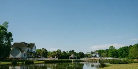 A scenic view of a lakefront wedding venue featuring a white bridge, a gazebo, and houses surrounded by green lawns and trees.