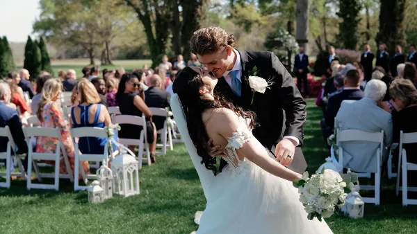 A bride and groom kiss during an outdoor wedding ceremony with guests seated in white chairs.