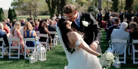 A bride and groom kiss during an outdoor wedding ceremony with guests seated in white chairs.