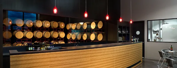 Interior view of a modern winery tasting room featuring a long wooden bar, wine bottles, and rows of oak barrels displayed on red racks behind the counter.