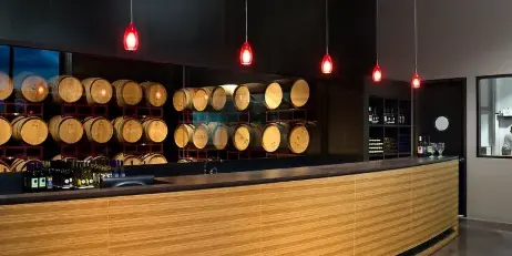 Interior view of a modern winery tasting room featuring a long wooden bar, wine bottles, and rows of oak barrels displayed on red racks behind the counter.
