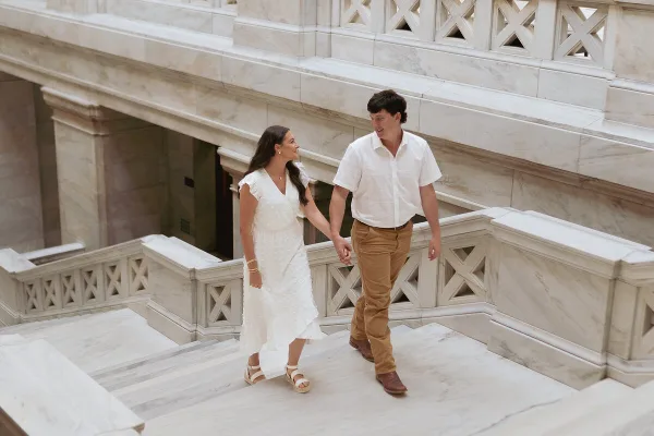 A couple holding hands and walking down a marble staircase in a grand building.