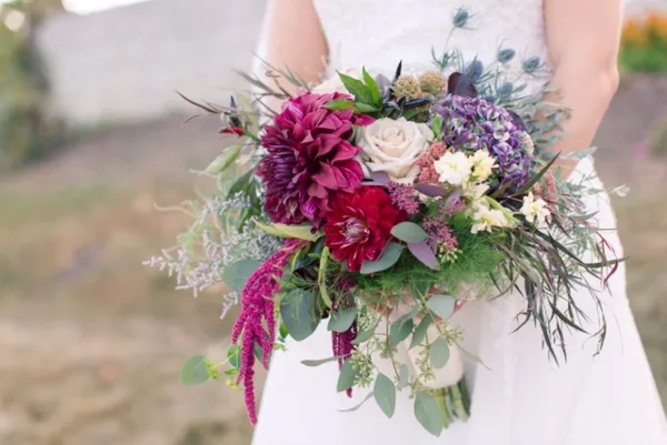 A bride in a white dress holding a vibrant bouquet of red dahlias, white roses, and greenery.