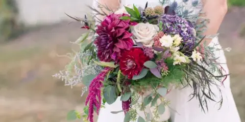 A bride in a white dress holding a vibrant bouquet of red dahlias, white roses, and greenery.