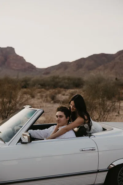A couple posing in a white convertible car with a desert mountain landscape in the background.