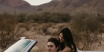 A couple posing in a white convertible car with a desert mountain landscape in the background.