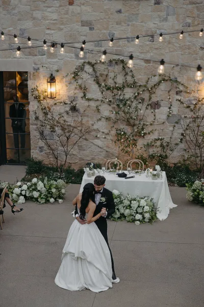 A bride and groom dancing at a wedding reception in front of a floral-decorated head table and stone wall.