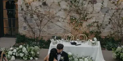 A bride and groom dancing at a wedding reception in front of a floral-decorated head table and stone wall.