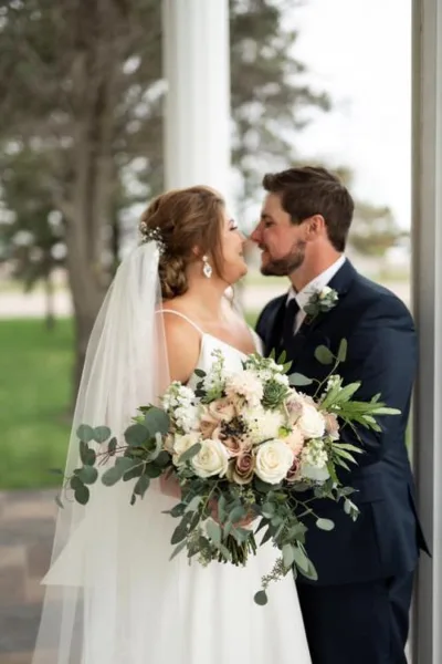 A bride and groom standing together, with the bride holding a large bouquet of white and blush roses with eucalyptus greenery and the groom wearing a matching boutonniere.