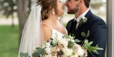 A bride and groom standing together, with the bride holding a large bouquet of white and blush roses with eucalyptus greenery and the groom wearing a matching boutonniere.