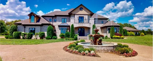 Exterior view of a large stone and stucco estate house with a fountain in the front yard.