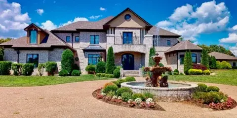 Exterior view of a large stone and stucco estate house with a fountain in the front yard.