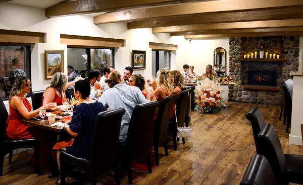 Interior view of a dining room with wood beams and a stone fireplace, showing guests seated at a long table during an event.